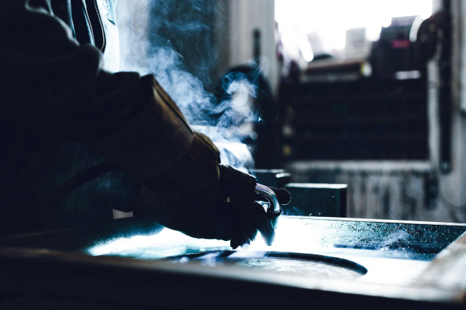 Welder working with intense arc light and rising smoke in a dimly lit workshop, focusing on metal fabrication.